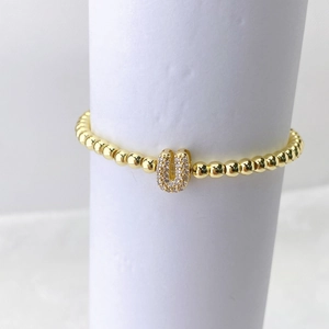 Detalle de pulsera elástica con cuentas doradas y un dije de letra U con circonitas incrustadas