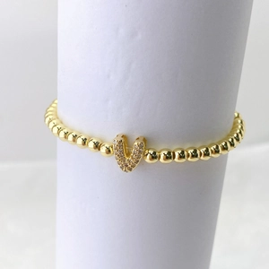 Detalle de una pulsera de cuentas de latón dorado con colgante de letra V y pavé de circón