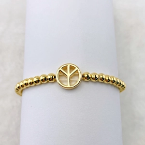 Detail eines goldenen Perlenarmbands mit einem runden Peace-Zeichen-Anhänger