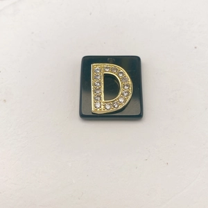 Detailansicht eines schwarzen quadratischen Anhängers mit goldenem Buchstaben D und Kristallen