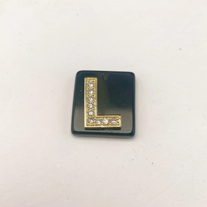 Detail eines quadratischen schwarzen Anhängers mit einem goldener Buchstabe L und Kristallen