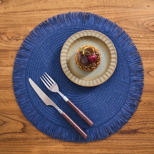 Top-down view of a navy blue woven paper placemat with delicate fringe on wood background