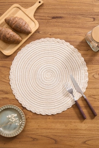 Top-down view of a round beige woven placemat with wavy lace edges on dark wood