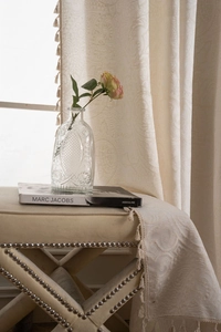 Detail of cream damascus jacquard curtain fabric with elegant tassel trim in soft sunlight