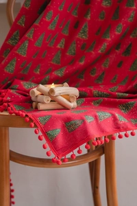 Close-up of red Christmas fabric with green tree patterns and pom-pom trim on wood
