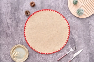 Top view of a round beige woven placemat with red scalloped edges on a grey surface