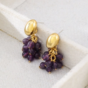 Elegantes pendientes dorados con racimos de uvas moradas sobre un joyero blanco
