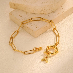 Detail van een gouden paperclip-kettingarmband met een pavé-letter A-bedel op een stenen achtergrond