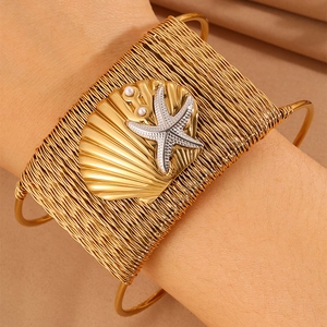 Macro detail of a wide woven gold bangle with a shell, starfish, and pearls on wrist