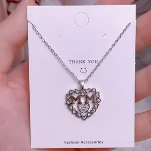 Detalle del collar MOM con forma de corazón plateado y cristales en tarjeta blanca