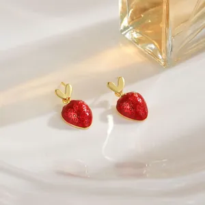 Detail of red strawberry earrings with golden heart leaves on a wavy white surface