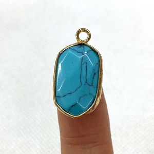 Faceted turquoise stone pendant with gold trim held on fingertips over white fabric
