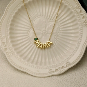 Close-up of gold MAMA necklace with a green teardrop gemstone on a ceramic tray