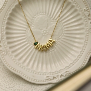 Close-up of golden MAMA necklace with green birthstone detail on a white ceramic tray