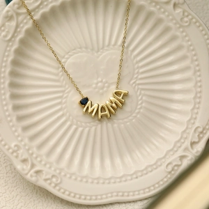 Detailed close-up of gold MAMA necklace with a blue teardrop gemstone on a ceramic plate