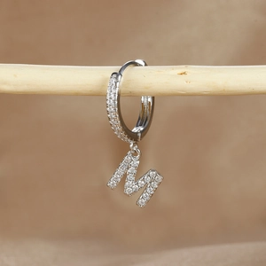 Detailed view of silver hoop earring with crystal-encrusted letter M pendant on wood
