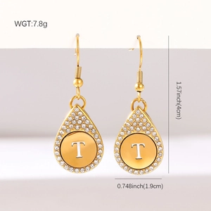 Détail de boucles d'oreilles dorées en goutte avec lettre T et halo de cristaux sur support