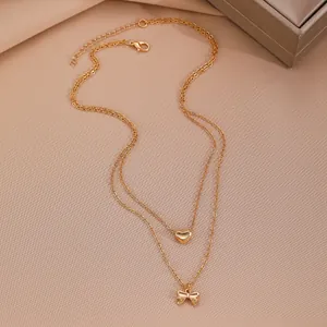 Detail of double-layer gold necklace featuring heart and bow pendants on a warm beige background