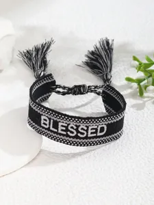 Detalle de pulsera tejida en blanco y negro con texto BLESSED bordado y adornos de borlas