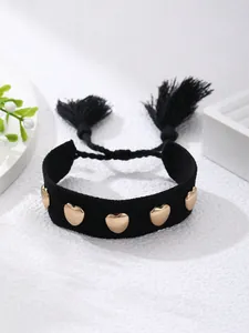 Detalle de pulsera tejida negra con tachuelas de corazón dorado y borlas en fondo blanco