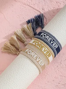 Detalle macro de tres pulseras tejidas con borlas y letras Forever sobre algodón
