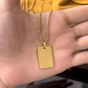 Dettaglio della collana a sfere dorata con pendente quadrato inciso Breathe tenuto in mano