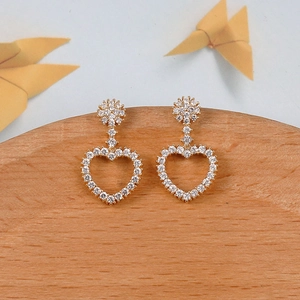 Detalle de pendientes de corazón dorados con pavé de piedras blancas y diseño floral de margarita