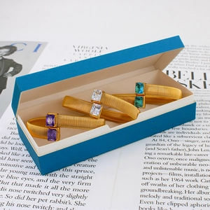 Detail van drie gouden bypass-armbanden met rechthoekige edelstenen in een blauwe doos