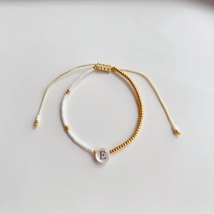Detail van verstelbare kralenarmband met witte vierkante kralen en gouden letter E bedel