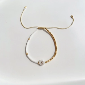 Detail van een kralenarmband met witte en gouden kralen en een S-letterbedel