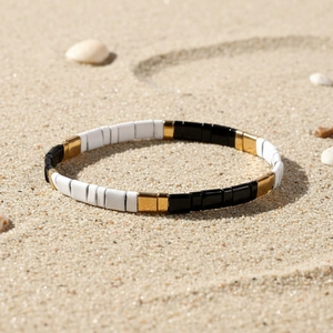 Dettaglio del braccialetto di perline Tila nero, bianco e oro sulla sabbia della spiaggia