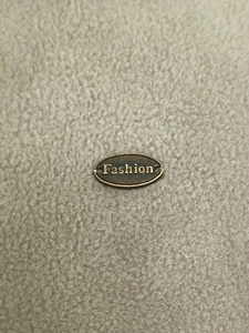 Detail of a bronze oval metal tag with Fashion engraving on gray fleece sweatshirt fabric