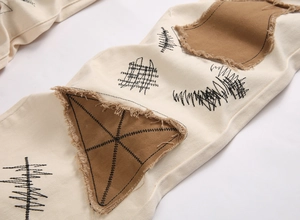 Detailed close-up of distressed brown patches and black embroidery on off-white denim pants
