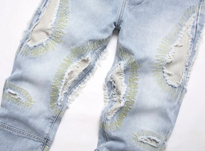 Detail of distressed light blue denim jeans with yellow zigzag embroidery and ripped patches
