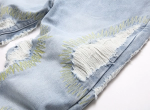 Close-up of light blue denim fabric with distressed rips and yellow wave embroidery details