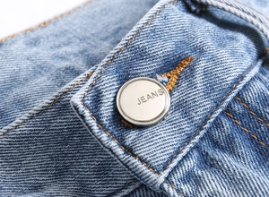 Détail macro du bouton argenté gravé JEANS sur le pantalon en jean bleu évasé