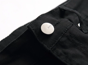 Macro shot of black denim waistband featuring a polished silver metal button engraved with JEANS