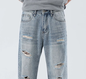 Detail of light blue washed denim jeans featuring distressed ripped holes and metal button