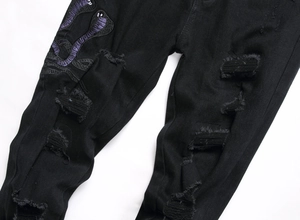 Detailansicht der lila Schlangenstickerei und starken Rissen auf schwarzen Denim-Jeans