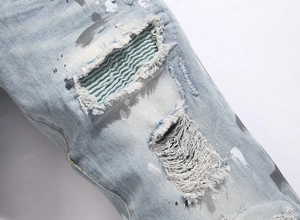 Detail of blue distressed jeans with frayed holes, fabric patches, and white paint splatters
