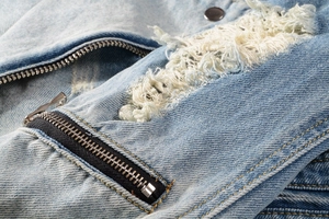 Macro detail of metal zipper and distressed hole design on light blue denim jacket