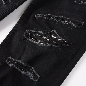 Macro detail of black denim jeans showing distressed holes and paisley fabric patch
