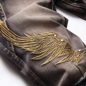 Men's distressed jeans featuring golden wing embroidery and metallic rivet details
