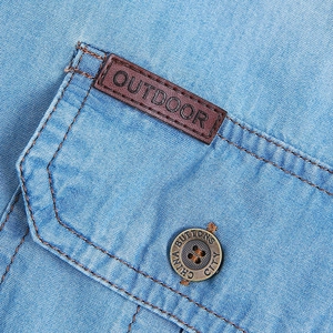 Detailed close-up of light blue denim shirt pocket with outdoor leather patch and metal button