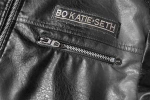 Detail einer schwarzen PU-Lederjacke mit geprägtem Marken-Patch und Metall-Reißverschlusstasche