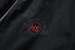 Detail of a black jacket featuring a red HS Hunter Story circular embroidery and metal zipper