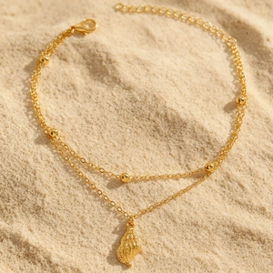 Nahaufnahme einer goldenen Boho-Fußkette mit Muschel-Anhänger auf echtem Sandstrand