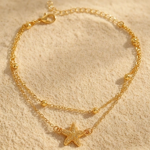 Detailansicht einer goldenen zweilagigen Fußkette mit Seestern-Anhänger auf Sand.
