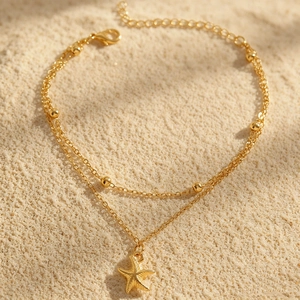 Detailansicht eines goldenen zweilagigen Fußkettchens mit Seestern-Anhänger auf Sand.