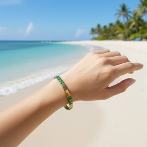 Bracelet à perles vert et doré porté au poignet sur un fond de plage ensoleillée tropicale